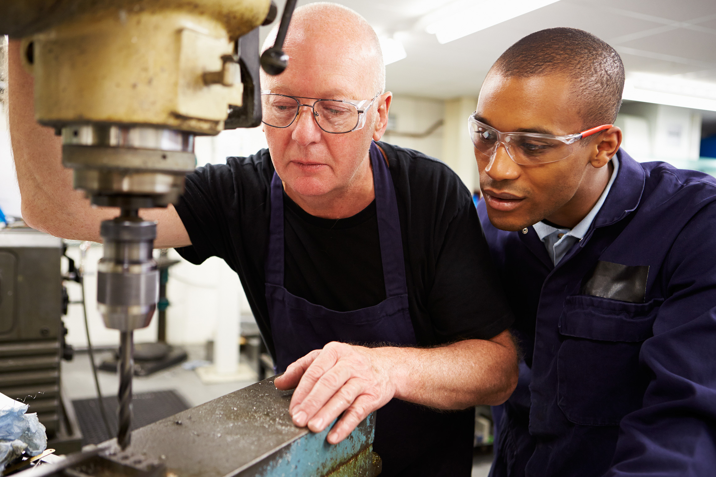 a younger man watches as an older man uses a drill machine.