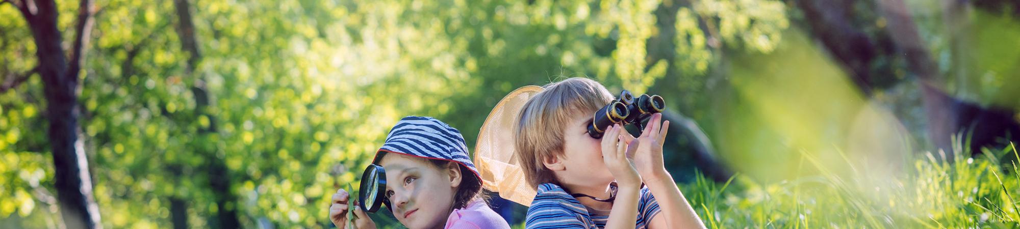 two children sitting in a wooded area looking through magnifiers at their surroundings